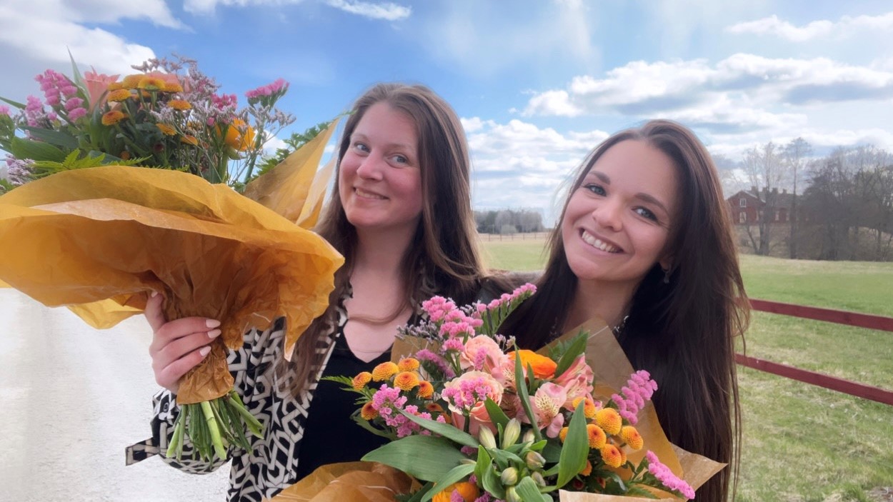 Emma Green och Emma Langgren är Årets Unga Företagare 2022 i Heby kommun. Här står de med varsin blombukett i lantlig miljö.