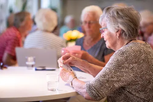 Äldre dam spelar kort vid ett bord. I bakgrunden fler personer. 