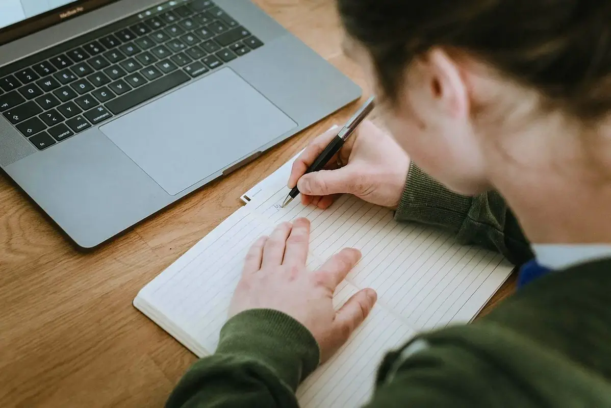 Kvinna skriver i anteckningsbok. På bordet står en bärbar dator. 