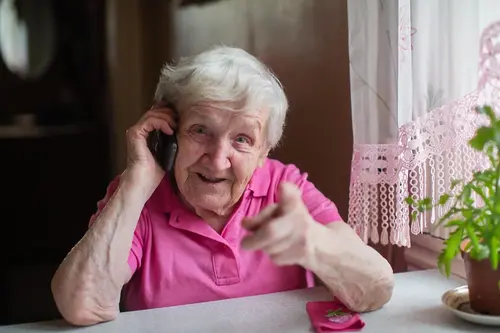 Senior kvinna sitter vid bord och pratar i telefon, samtidigt som hon pekar glatt mot fotografen