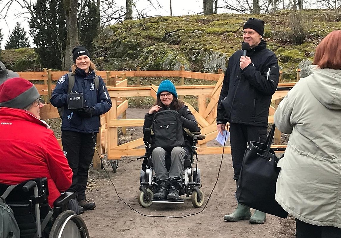 Landshövning Göran Enander invigningstalar framför grinden till Aspnäs naturreservat. Bredvid honom rullstolsburna Karin Nordström. 